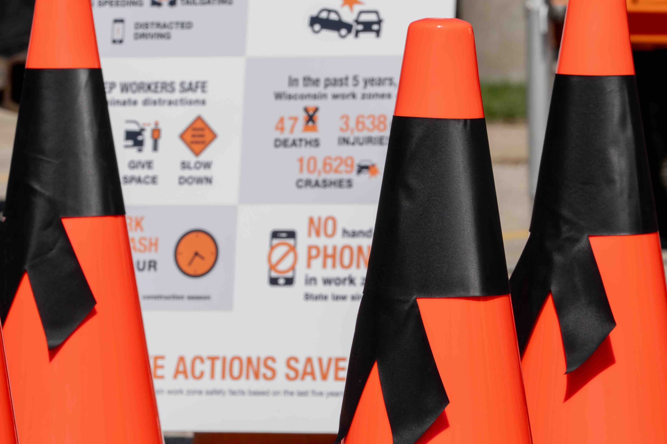 Orange and black safety cones in the foreground with a blurred safety infographic/signage in the background.