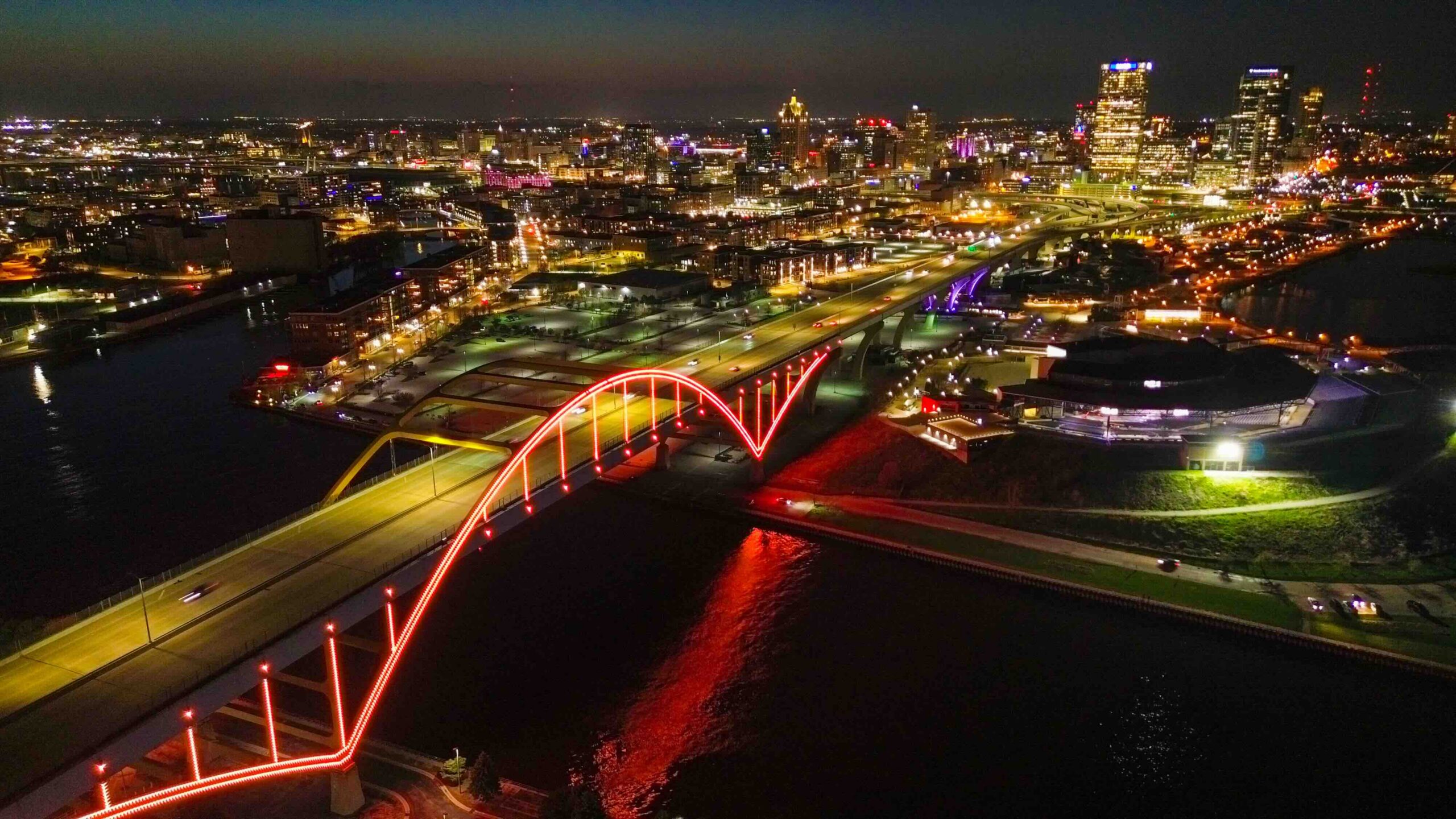 Aerial night view of a city skyline with a bright red illuminated arch bridge crossing a river.