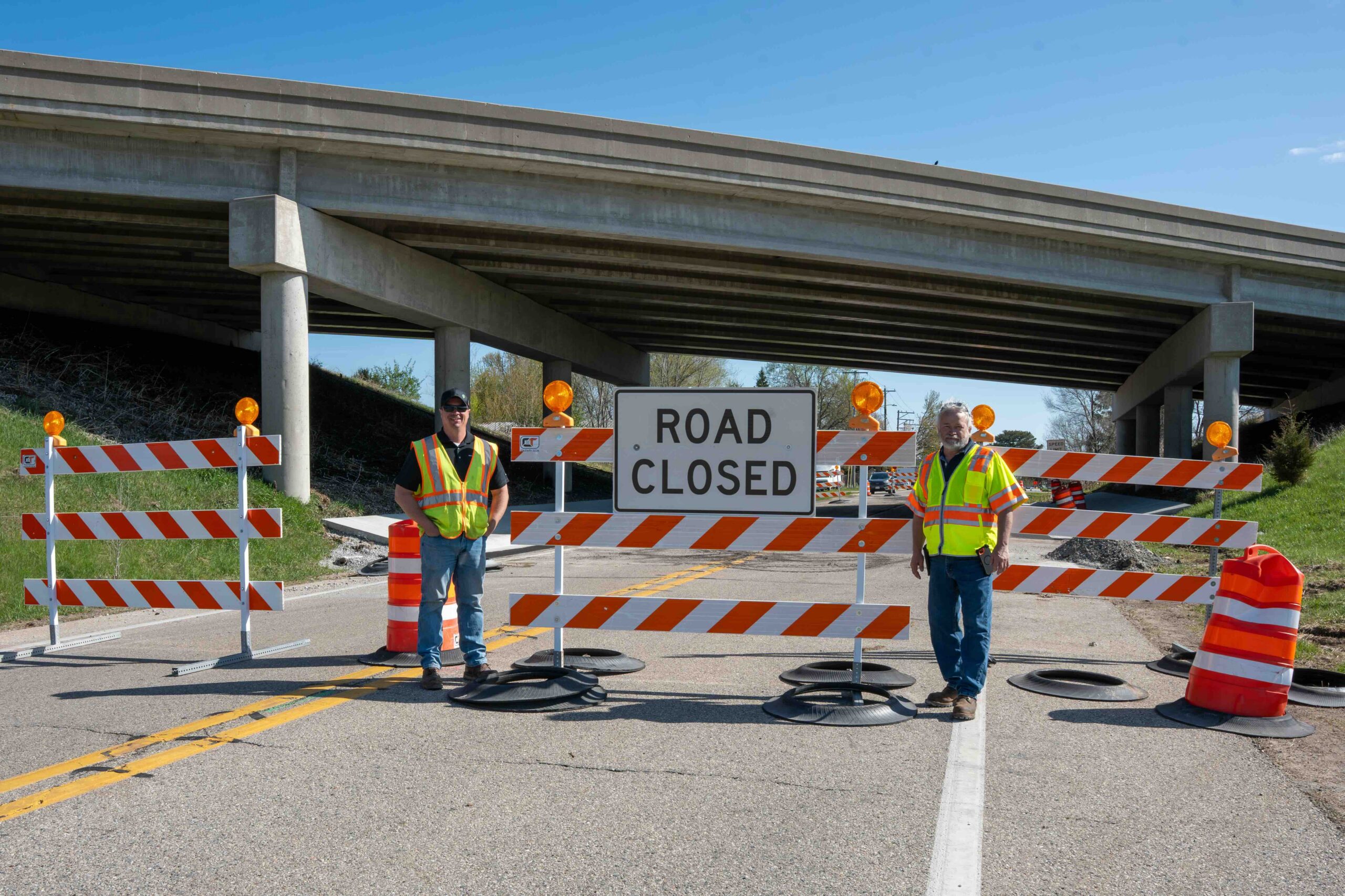 Two construction workers in high-visibility vests stand behind orange barriers blocking a closed road under an overpass.