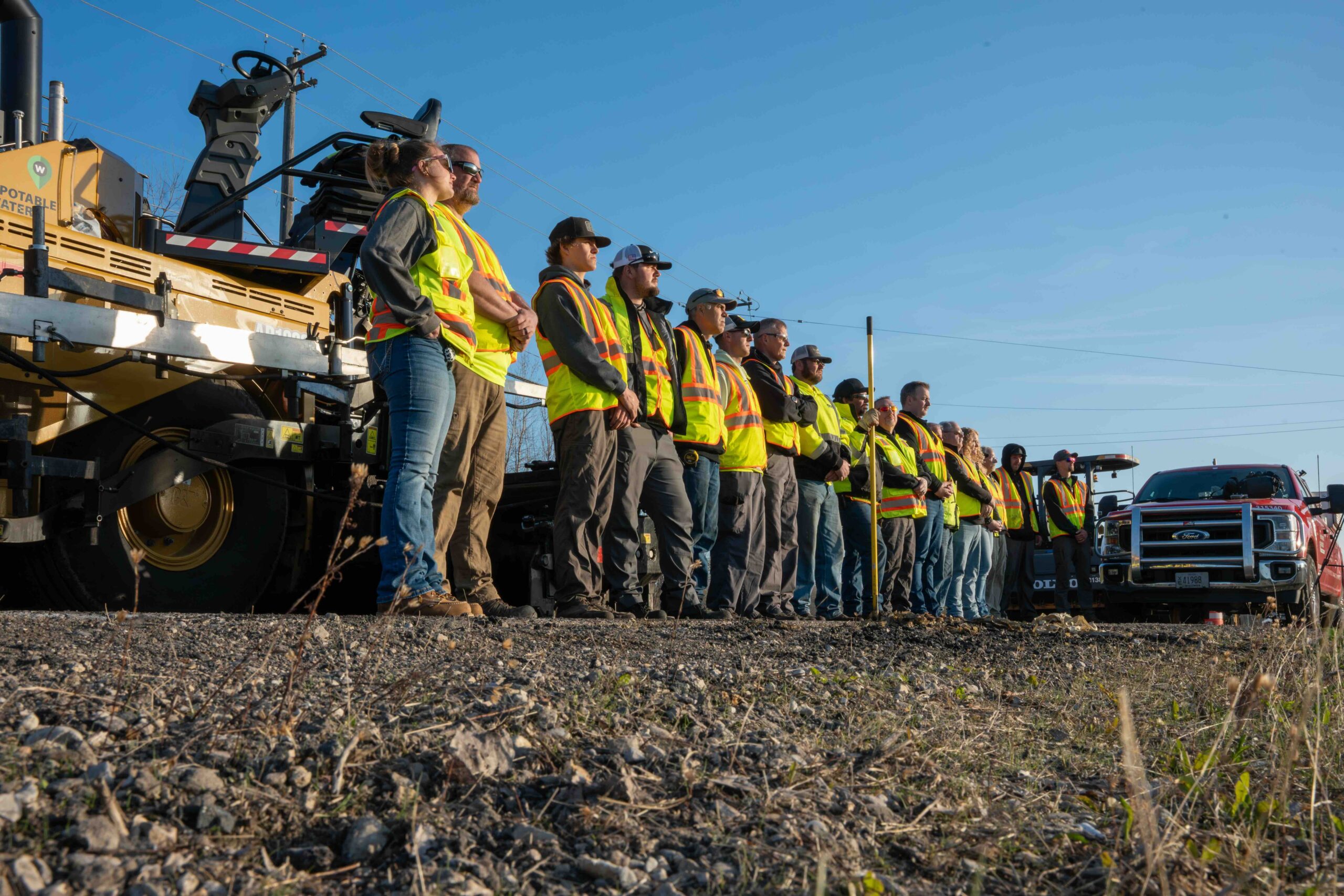 Group of construction workers in high-visibility vests standing on a platform beside a large yellow drilling machine, under a clear blue sky.
