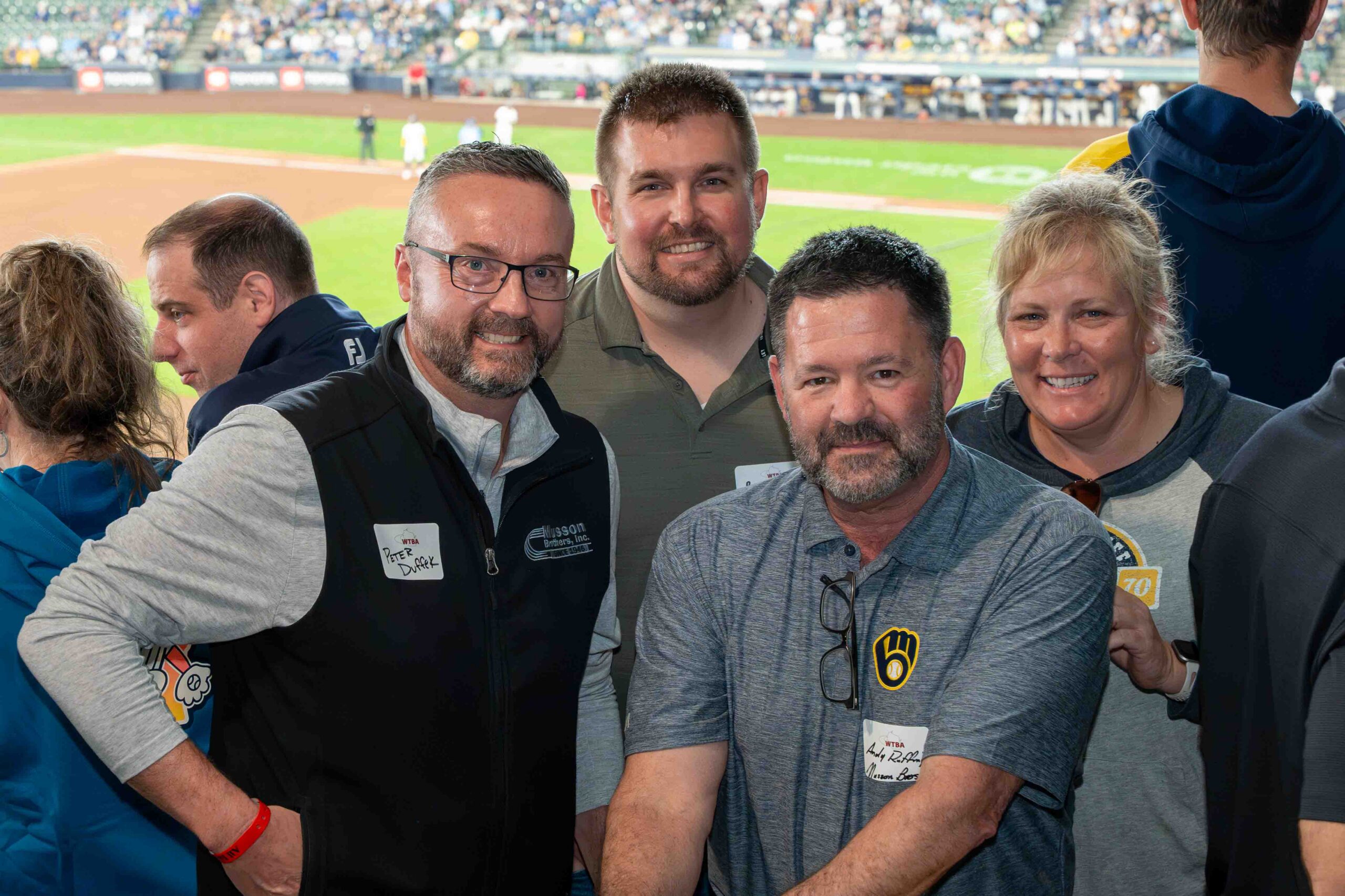 Five adults pose for a group photo in the stands with a baseball field in the background, all smiling at the camera.