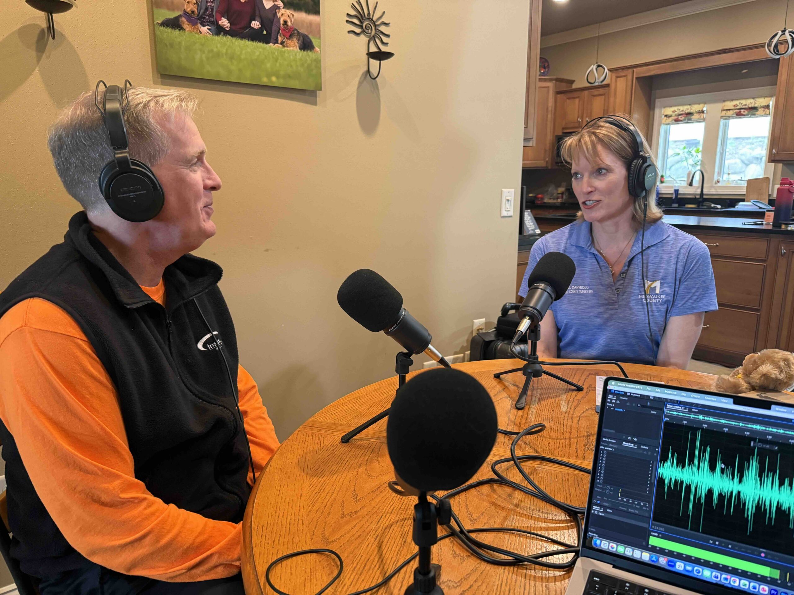 Two people wearing headphones sit at a round table with multiple microphones, recording a podcast in a home kitchen studio.