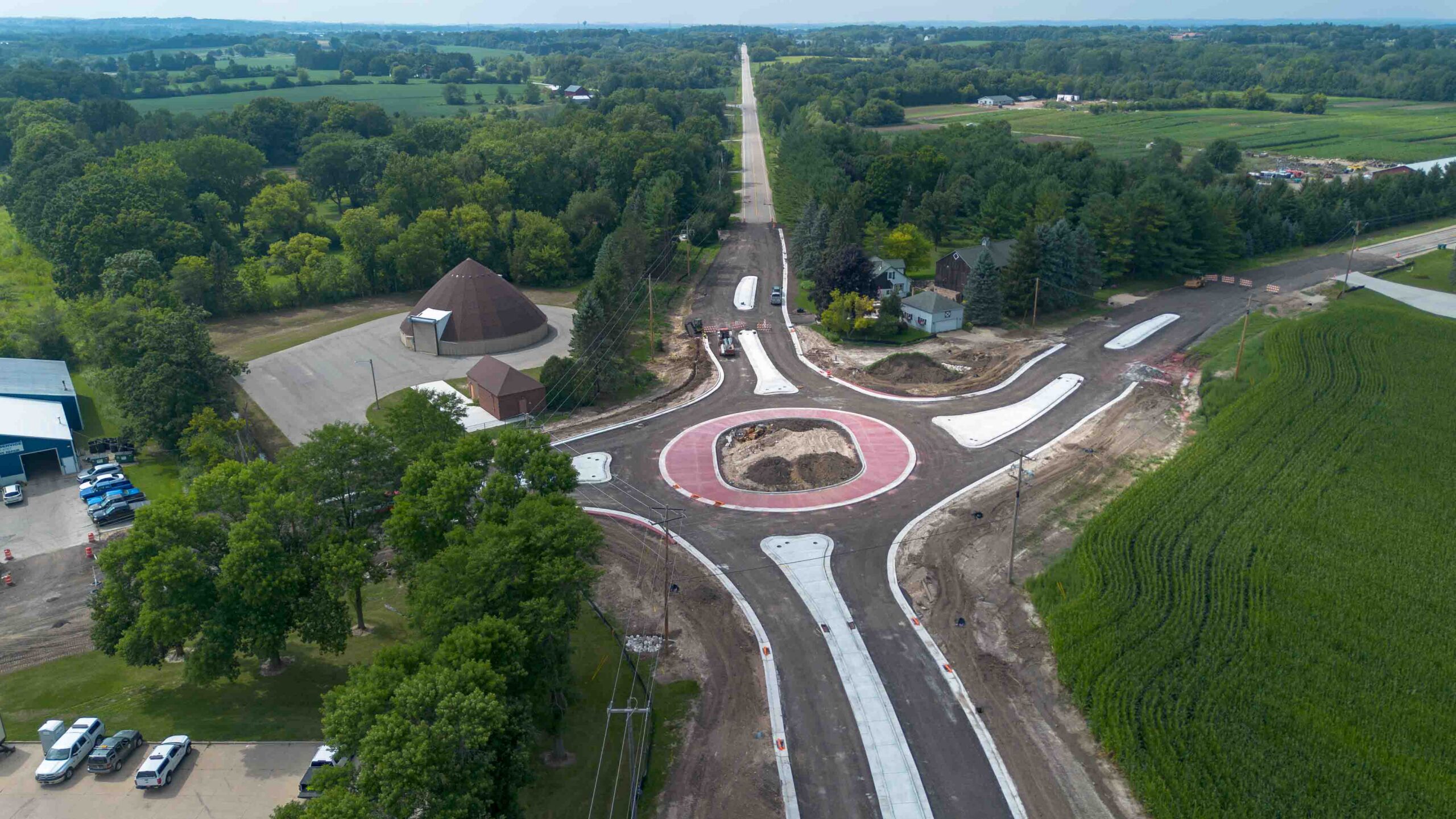 Aerial view of a road construction site with a looping roundabout, newly paved lanes, and a red circular island, surrounded by trees and fields.