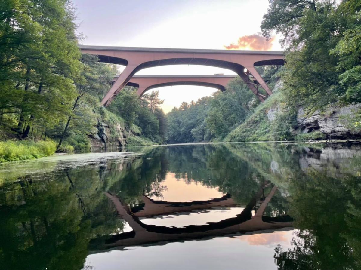 Arched brown bridge over a calm, tree-lined river with a clear reflection in the water.