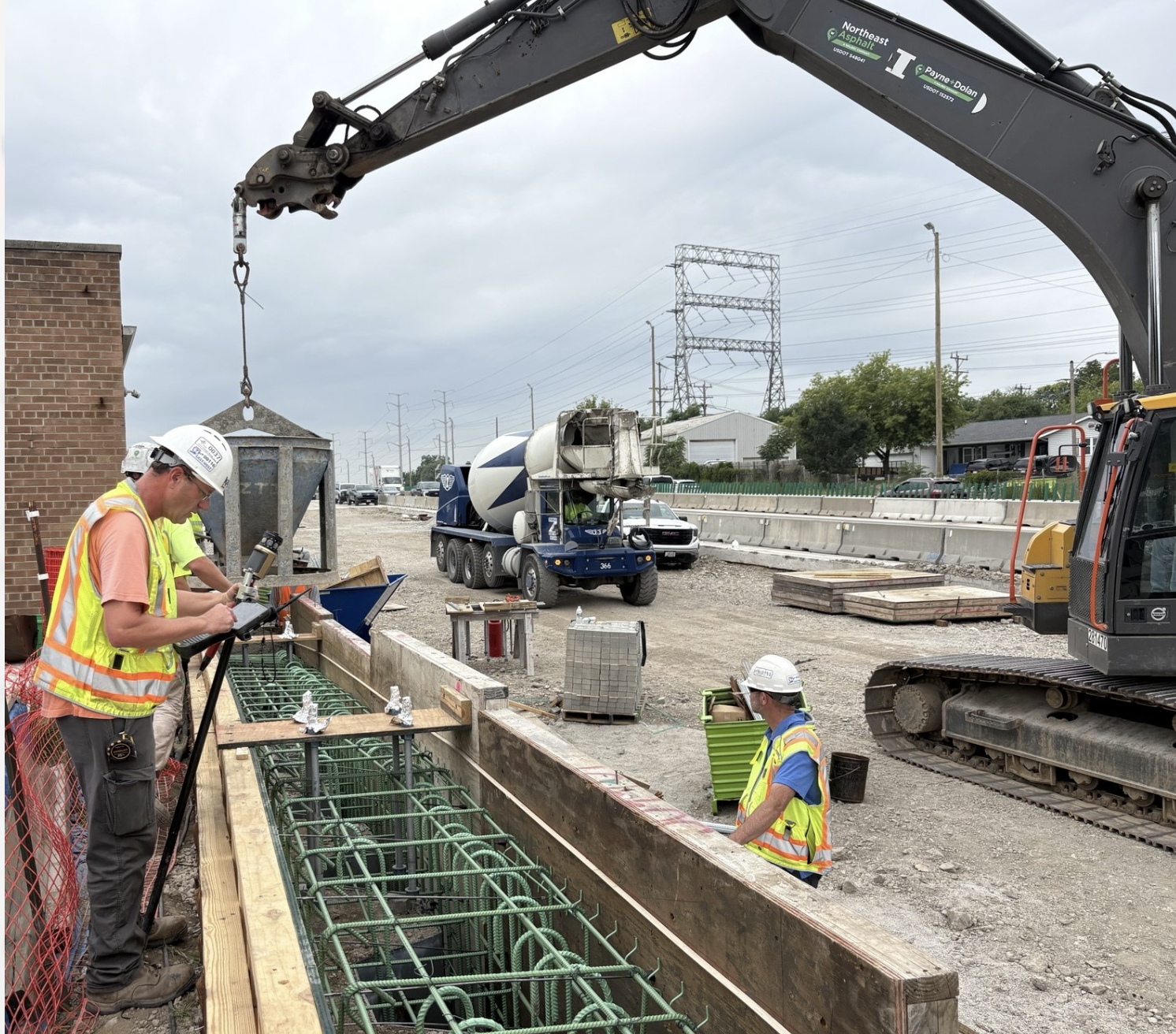 Road construction site: workers in safety vests and hard hats stand by a trench with exposed green rebar while a large excavator bucket lowers into the pit.