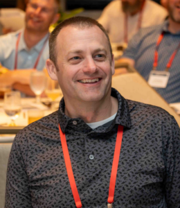 Smiling man at a conference, wearing a dark patterned shirt and a red lanyard, seated with other attendees in the background.