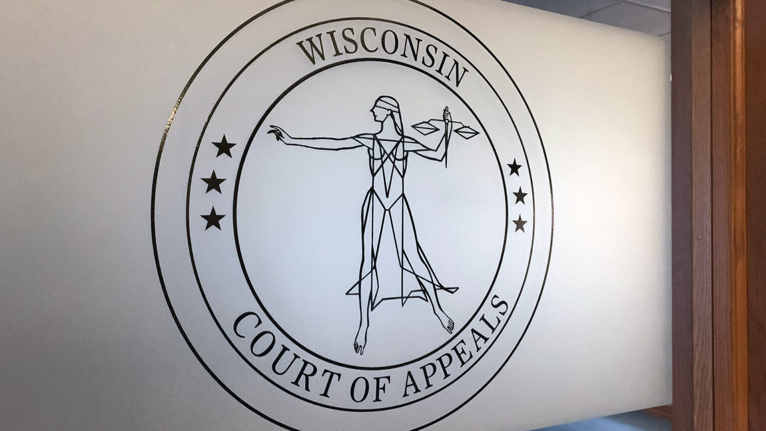 Circular Wisconsin Court of Appeals seal on frosted glass, featuring a stylized Lady Justice holding scales and a sword, with stars along the ring.